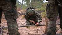 Ein Soldat hockt am Boden im australischen Busch und hat einen langen Stock quer vor sich zu liegen.