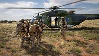 Vier Soldaten tragen eine verwundete Person auf einer Trage zum Hubschrauber. Weitere Soldaten sichern die Umgebung. 
