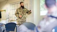 Ein Soldat steht an der Wand vor Lagekarten und hält eine Präsentation. Andere Soldaten folgen seinen Ausführungen. 