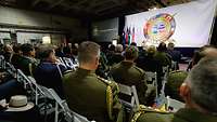 Soldaten sitzen in einem Saal und lauschen einem Redner vor einer weißen Leinwand mit Emblem.