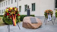 Ein Stein mit Bronzetafel auf dem Henning und Erika von Tresckow 20.07.1944 steht, liegt zwischen 2 Blumenkränzen.