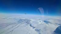 Die vereiste Landschaft von Island ist zu sehen. Im Horizont erkennt man die Erdkrümmung. Ganz klein in der Ferne ist ein Jet.