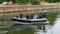 Ein Schlauchboot mit mehreren Soldaten fährt über ein Gewässer. Ein Soldat hält ein Schild hoch.