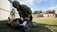 Ein verletzter Soldat liegt am Boden auf einer Wiese neben einem Zaun. Ein zweiter versorgt ihn.