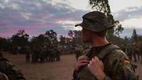 Ein Soldat steht abseits von Soldatengruppen, die draußen im Abendrot auf einer Wiese stehen.