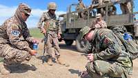 Zwei Soldaten hocken und einer steht vor einem Lkw, auf dem weitere Soldaten stehen.