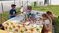 Vier Kinder sitzen an einem Tisch, darauf liegt eine bunte Tischdecke und malen. 