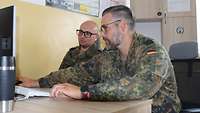 Zwei Soldaten sitzen an einem Schreibtisch mit einem Computer. Beide schauen auf den Bildschirm. 