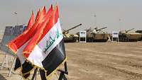 Im Vordergrund wehen irakische Nationalflaggen, während im Hintergrund verschiedene Kettenfahrzeuge zu sehen sind