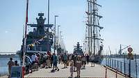 Gäste laufen auf einer Pier zwischen einem grauen Schiff und einem Segelschiff hindurch.