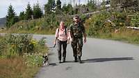 Ein Soldat marschiert gemeinsam mit einer Frau. Er trägt ein Kind auf dem Rücken. Ein Hund läuft auch mit.