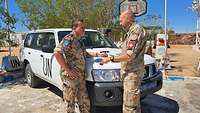 Zwei Soldaten verschiedener Herkunft stehen vor einem Auto und unterhalten sich, im Hintergrund ein Basketballkorb.