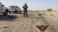 Ein Soldat mit blauem Helm auf einer Wüstenstraße macht sich Notizen, vorne ein Schild mit der Aufschrift „Danger Mines“. 