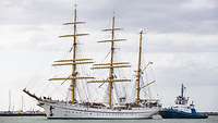 Das Segelschiff „Gorch Fock“ läuft in einem Hafen ein, daneben ist ein Begleitboot der Küstenwache.