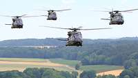 Vier Hubschrauber fliegen versetzt über eine Landschaft aus Bäumen und Feldern.