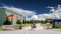 Blick auf die Zufahrt zu einer Kaserne mit Schlagbaum und Nationalflagge, dahinter links ein Neubaublock