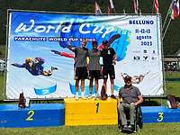 Drei Personen stehen auf einem Podium. Ein weiterer sitzt in einem Rollstuhl vor dem Podium.