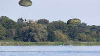 Zwei Fallschirmspringer kurz vor der Landung im Wasser. Im Hintergrund sind grüne Bäume.