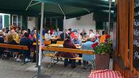 Mehrere Leute sitzen an Bierbänken und Tischen unter großen Schirmen im Biergarten.