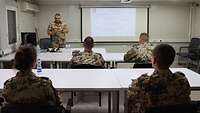 Vier Soldaten sitzen in einem Klassenraum an verschiedenen Tischen. Ein weiterer steht vor der Gruppe.