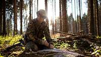 Ein Soldat hockt auf dem Waldboden und verbindet Äste und ein Metallgeflecht miteinander.