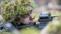 Ein Soldat liegt mit der Waffe im Anschlag im Wald in einer Stellung und beobachtet das Gelände.