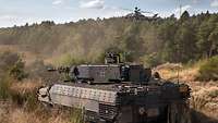 Ein Panzer steht im freien Gelände, im Hintergrund am Himmel ein Kampfhubschrauber Tiger.