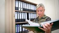 Eine Soldatin im Büro, die in einem Ordner liest und vor einem Regal mit Akten steht