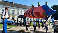 Ein Mann auf Stelzen und ein riesiger Fisch-Ballon vor einer Schlosskulisse