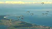 Fallschirmjäger gleiten am geöffneten Fallschirm gen Boden, unter ihnen die Küste und das Meer.
