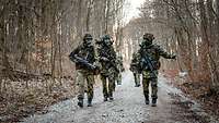 Mehrere Soldaten marschieren mit ABC-Maske und Ausrüstung durch einen Wald.