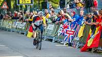 Sportler auf Rennrad fährt an Zuschauern vorbei, die mit Flaggen hinter einer Absperrung stehen