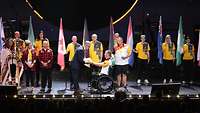 Im Rollstuhl sitzender deutscher Sportler übergibt Fahne an Mann in Anzug, im Hintergrund halten Frauen und Männer Flaggen