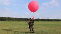 Ein Soldat steht mit einem roten Wetterballon auf einer Wiese. 