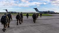 Soldaten mit Fallschirmen laufen über das Flugfeld zu ihrem Transportflugzeug.