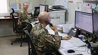 Zwei Soldaten sitzen nebeneinander an Schreibtischen vor ihren Computern. Ein Soldat lacht, der andere telefoniert.