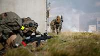 Ein Soldat liegt mit Waffe in der Hand an der Hausecke, vor ihm laufen Soldaten an der Wand entlang.