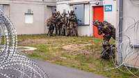 Soldaten stehen an einer Häuserecke und sichern mit ihren Waffen, im Vordergrund Stacheldraht.
