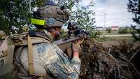 Ein Soldat im Wald blickt mit dem Gewehr im Anschlag aus einer Stellung auf eine Stadt.