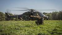 Ein Hubschrauber steht auf einer Wiese, Soldaten verlassen ihn, ein weiterer weist hockend den Weg.