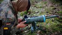 Ein Soldat kniet auf dem Waldboden und baut eine Sprengladung auf.