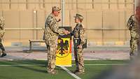 Eine Soldatin schüttelt einem Soldaten auf einem Fußballfeld die Hand, hinter ihnen ein Rednerpult mit Deutschlandflagge.