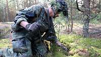 Ein Soldat kniet auf einem Waldboden und durchforstet mit einer Hand das Moos.
