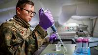 Ein Soldat mit einer Pipette und lilafarbenen Gummihandschuhen arbeitet im Labor in einem Zelt.