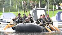 Soldaten paddeln mit Schlauchboot auf einem Fluss. Vorn liegt ein Soldat mit Maschinengewehr.