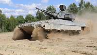 A tank moving from right to left through deep sand in front of a forest