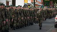 Soldatinnen und Soldaten marschieren in Formationen eine Straße entlang, im Hintergrund Häuser.