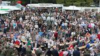 Ein Marktplatz voller Besucher, im Hintergrund ein Park, davor Getränke- und Grillwagen. 