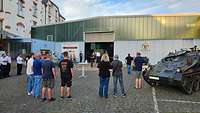 A group of people is standing in front of an exhibition hall. A tank is standing at the side.