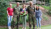 Eine Frau steht mit einem Blumenstrauß zwischen 2 Soldaten, daneben stehen zwei weitere Frauen. 